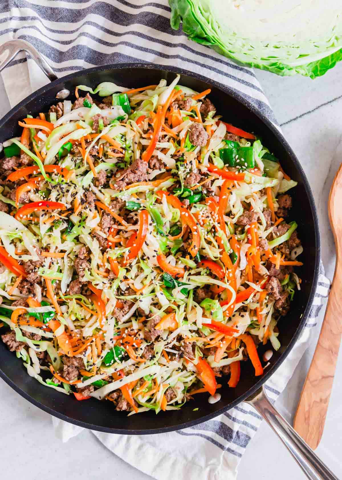 Overhead shot of ground beef and cabbage recipe in a black skillet with half a head of cabbage and wooden spoon on the side.