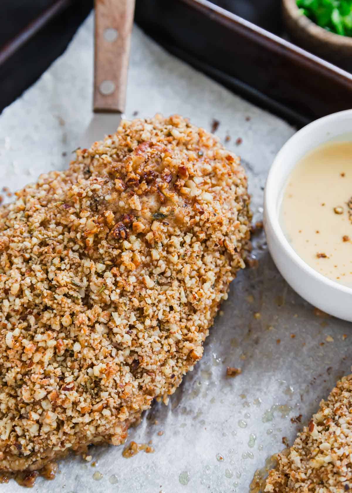 Pecan crusted chicken breast with a serving spatula on a baking sheet with a small bowl of honey dijon yogurt sauce on the side.