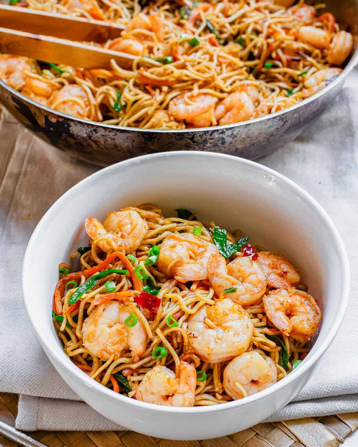 Shrimp lo mein in a white bowl with wok in the background.