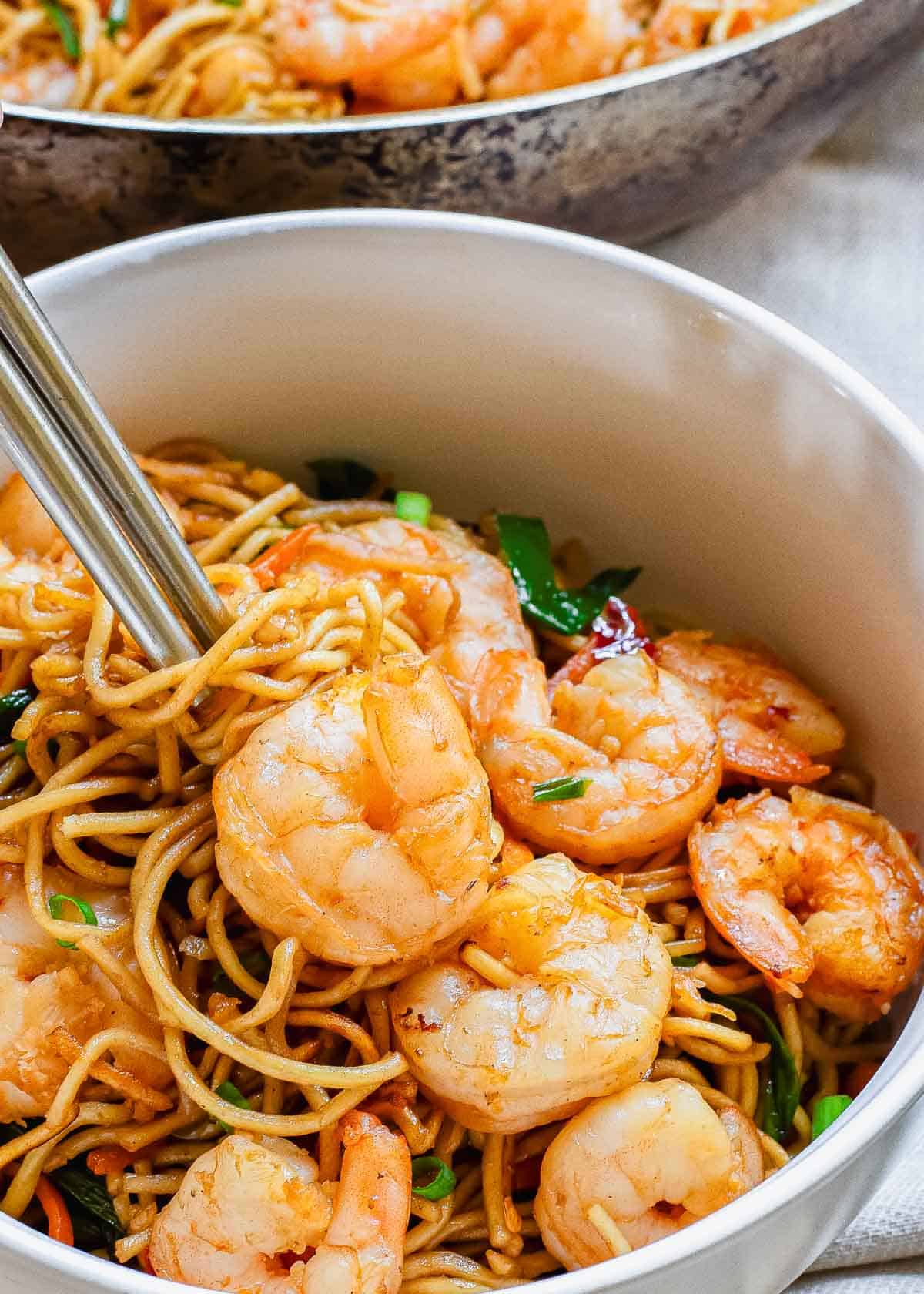 Quick shrimp lo mein in a white bowl with chopsticks and wok in the background.