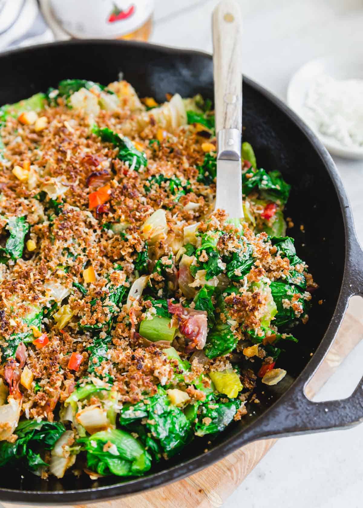 A skillet full of escarole with breadcrumbs and cheese on top with a serving spoon.