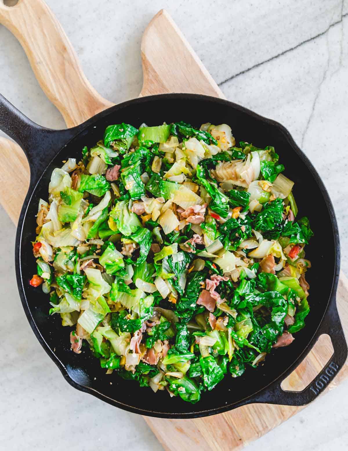 Escarole greens with onions, pickled hot peppers, prosciutto and garlic in a cast iron skillet on a wooden cutting board.