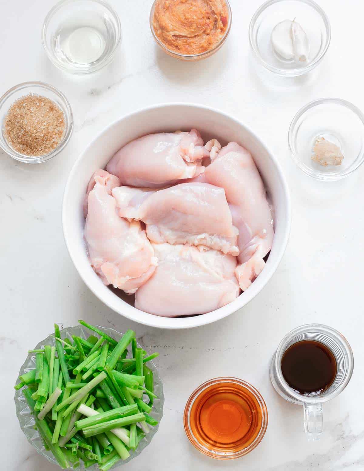 A bowl of raw chicken thighs surrounded by ingredients to make gochujang chicken.