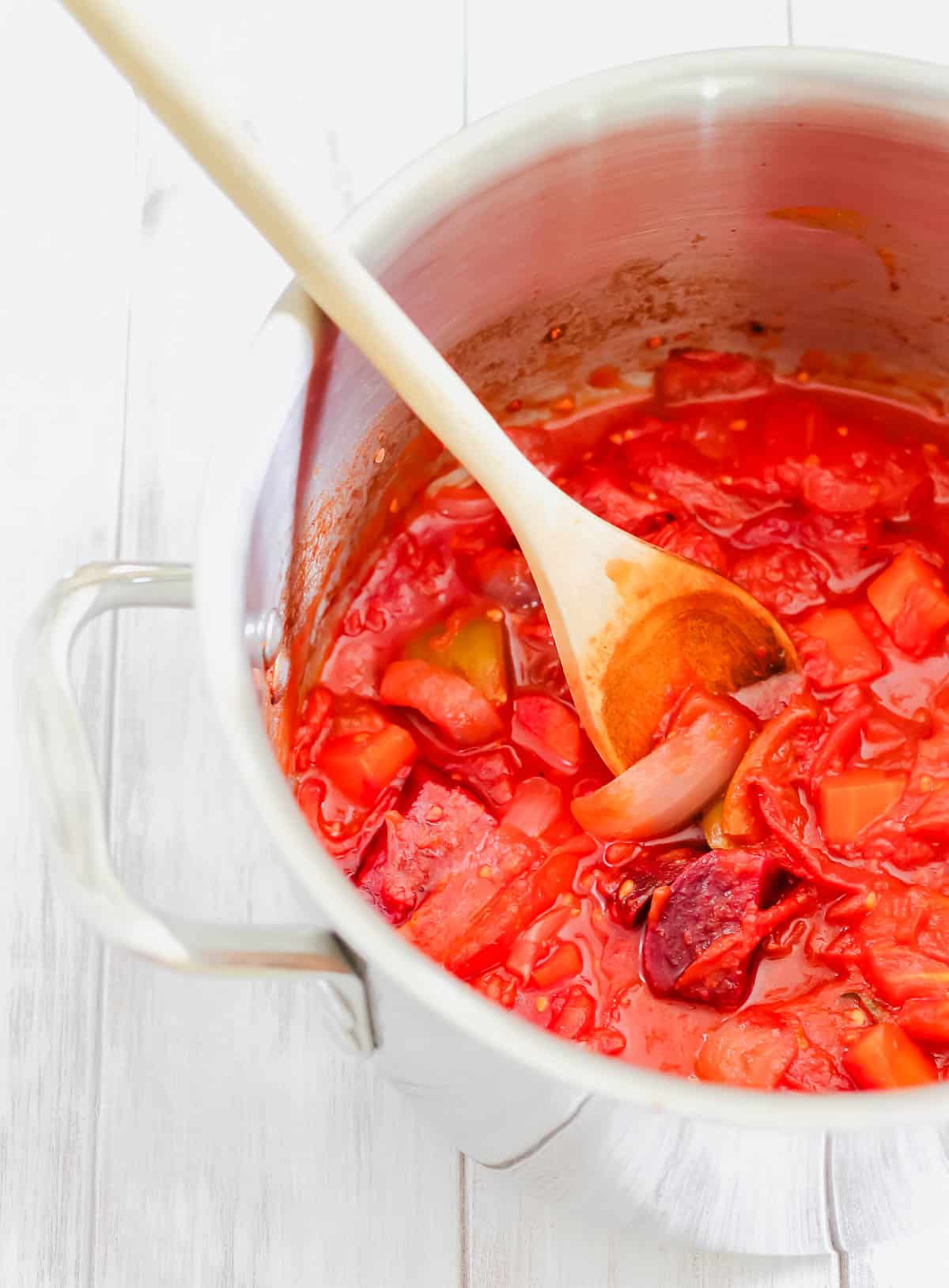 A pot of simmering tomatoes and vegetables with a wooden spoon.