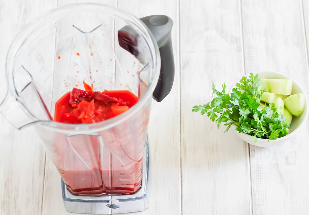 A blender filled with cooked vegetables and tomatoes with a small bowl of cucumbers and parsley on the side.