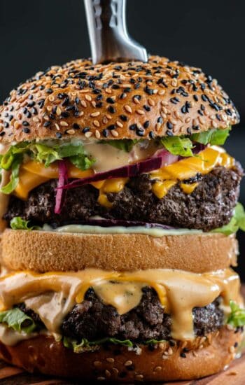 A double decker burger with cheese, lettuce, red onion and burger sauce on black and white sesame seed bun with a knife in the center.