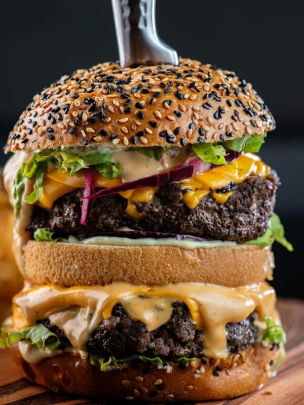 A double decker burger with cheese, lettuce, red onion and burger sauce on black and white sesame seed bun with a knife in the center.