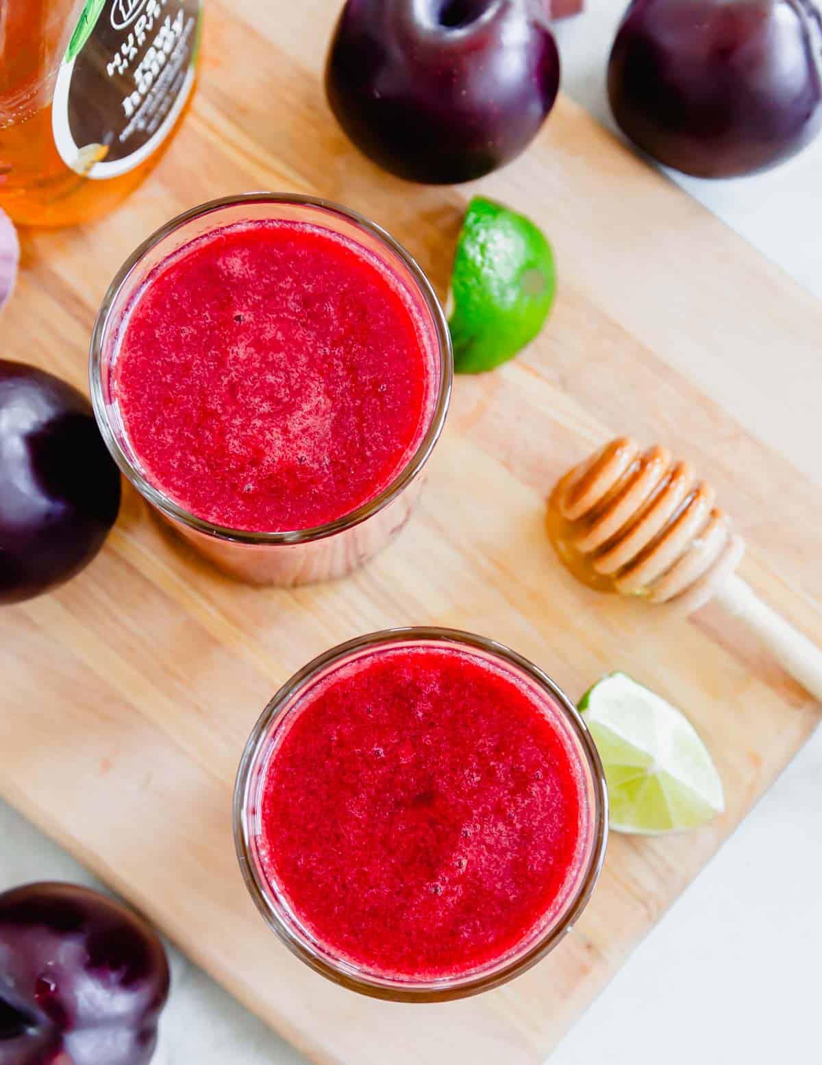 Overhead shot of two glasses of plum juice on a cutting board with lime wedges and honey.