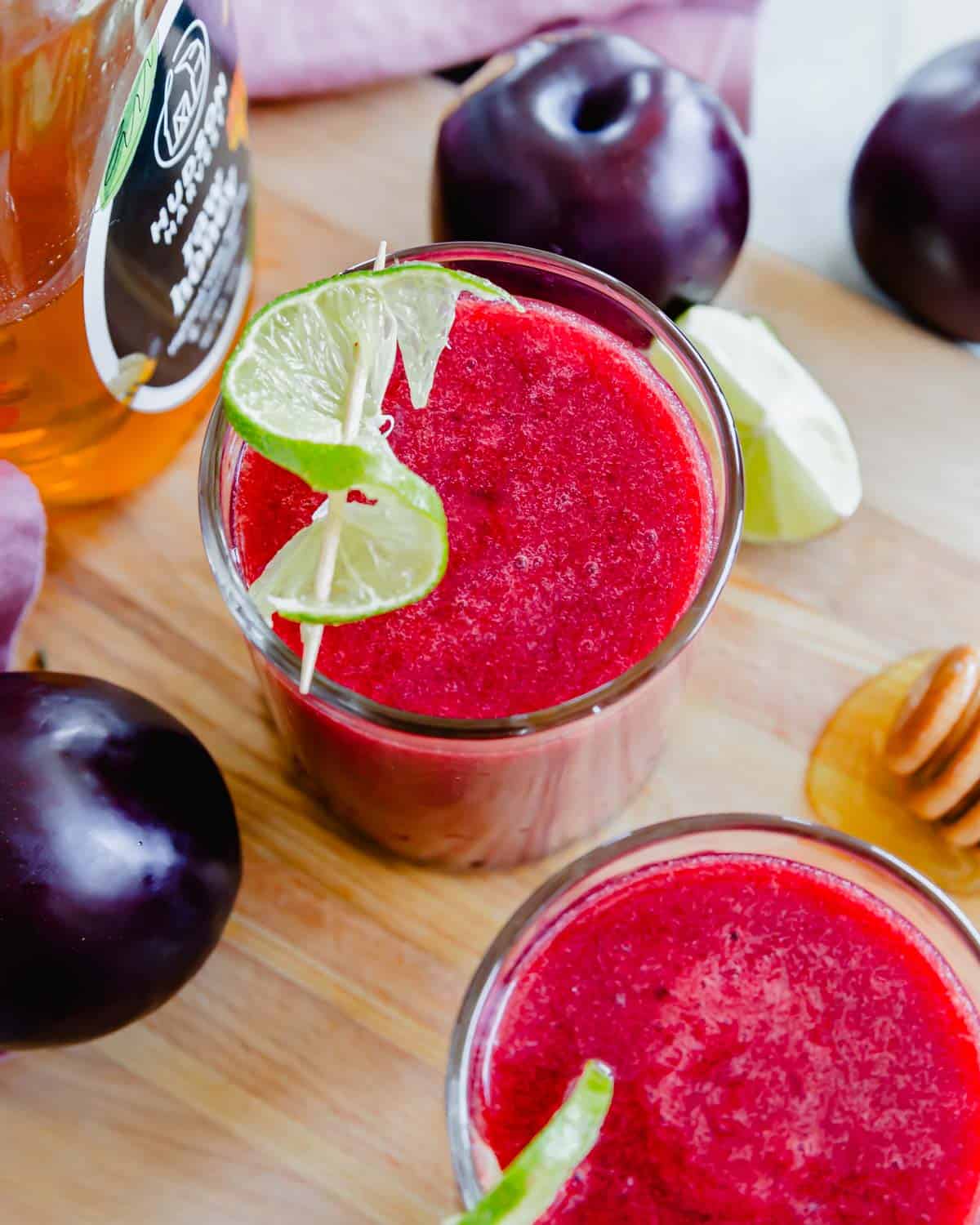 Two glasses of plum juice with lime garnish and honey on a cutting board.