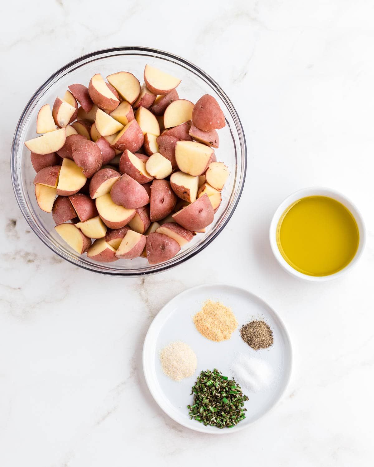 Red potatoes cut into small pieces in a glass bowl with oil and spices on the side.