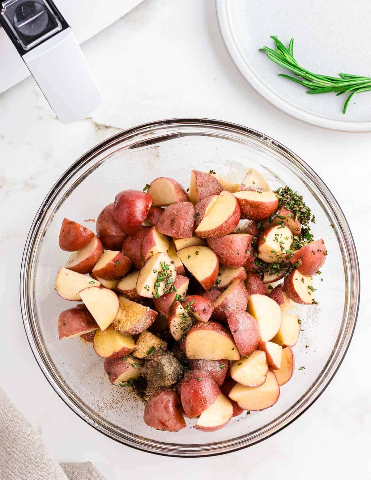 Red potatoes cut into chunks with fresh minced rosemary and spices in a glass bowl.