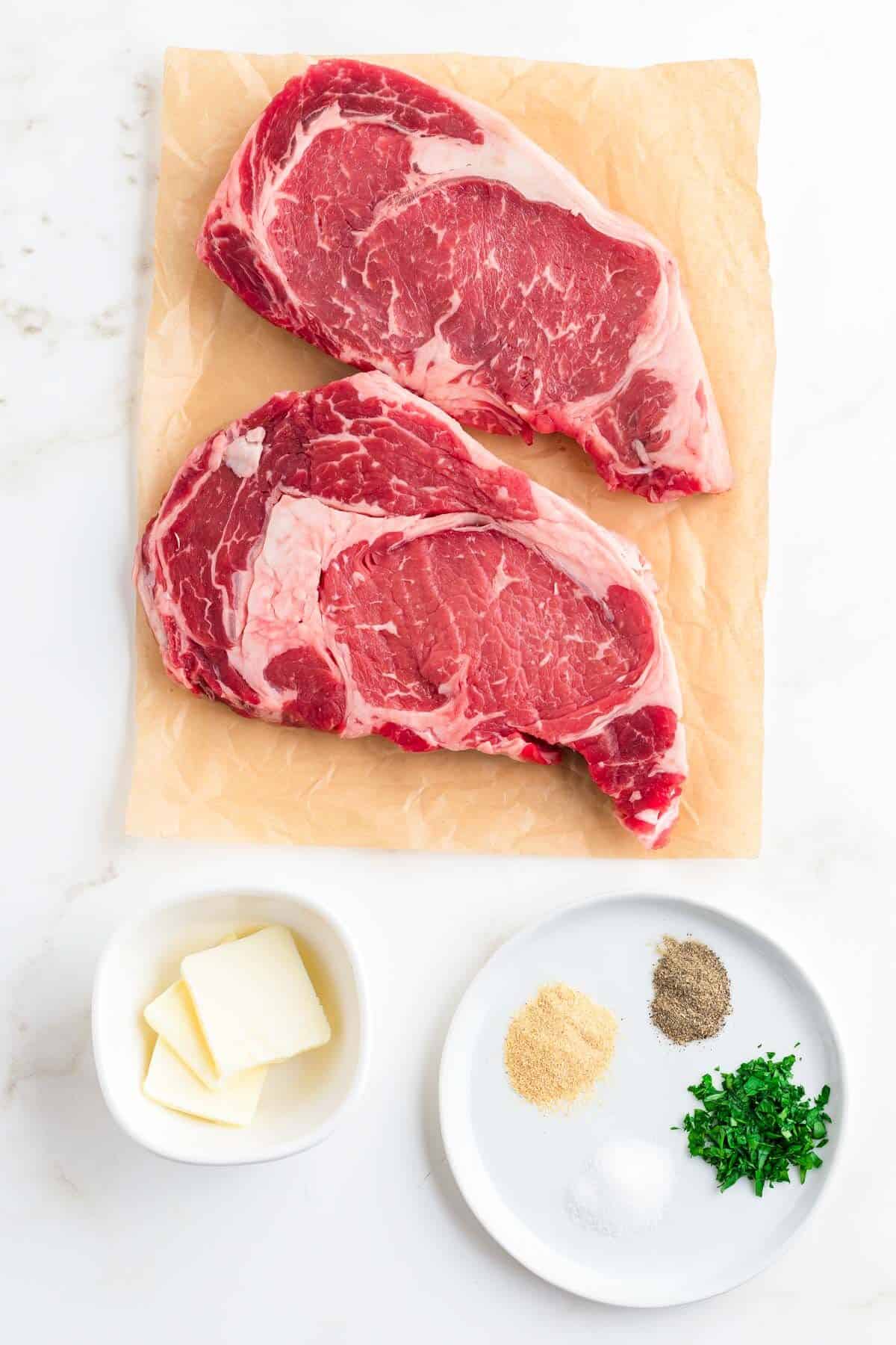 Two raw ribeye steaks on parchment paper with seasoning and butter in small bowls to the side.