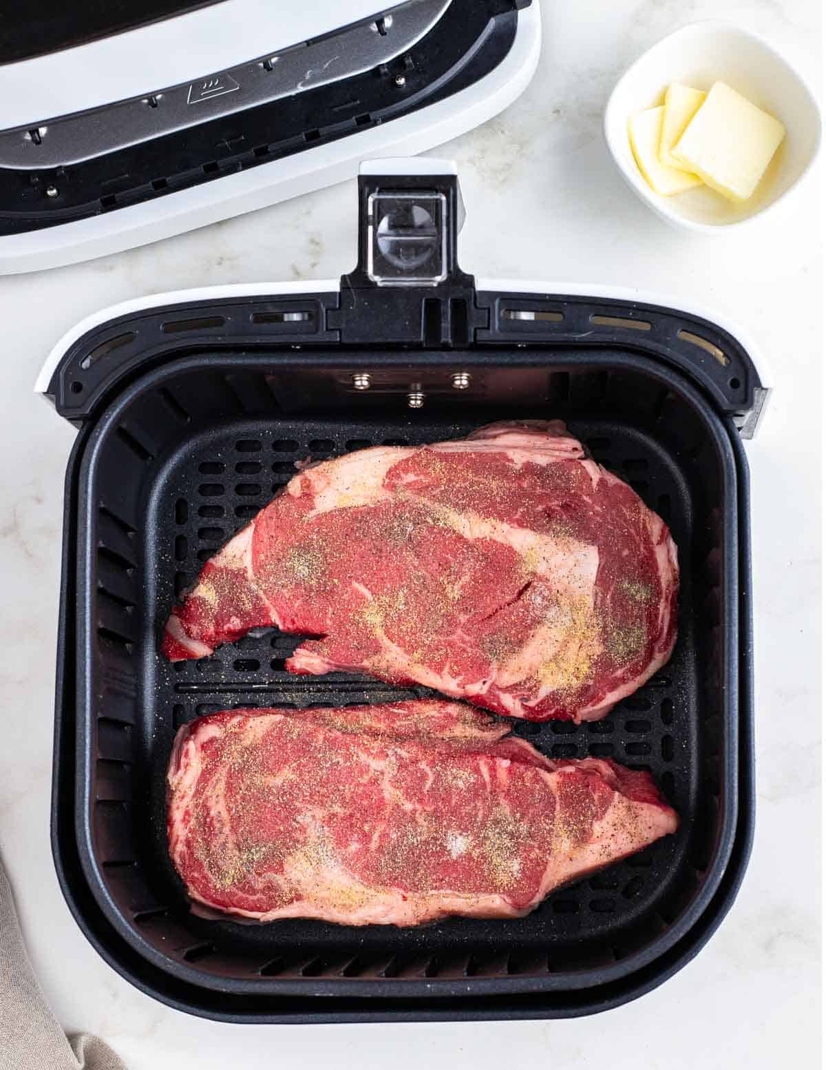 Seasoned ribeye steaks in an air fryer basket.