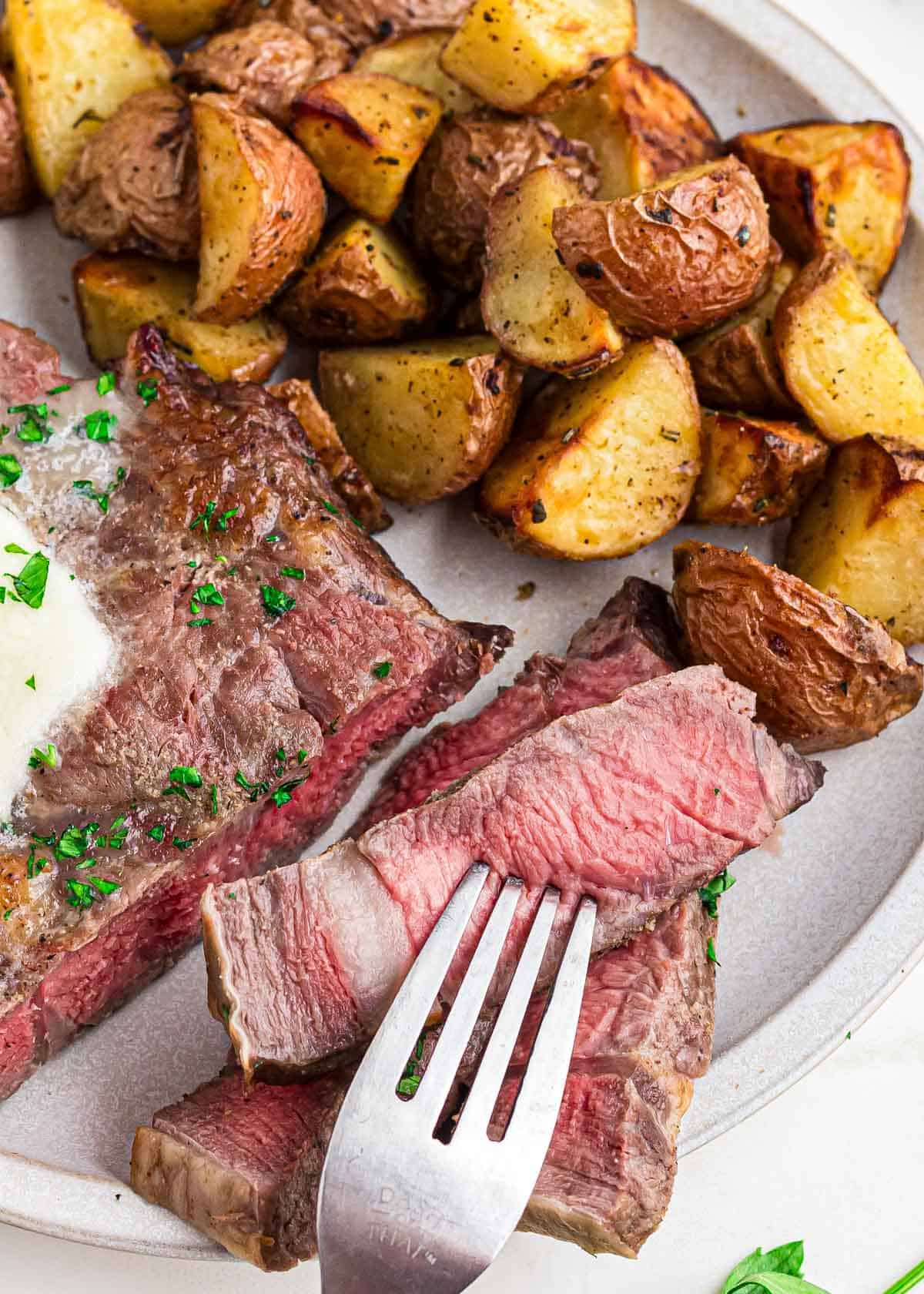 Close up of a fork in a piece of medium-rare cooked air fried ribeye steak on a plate with roasted potatoes.