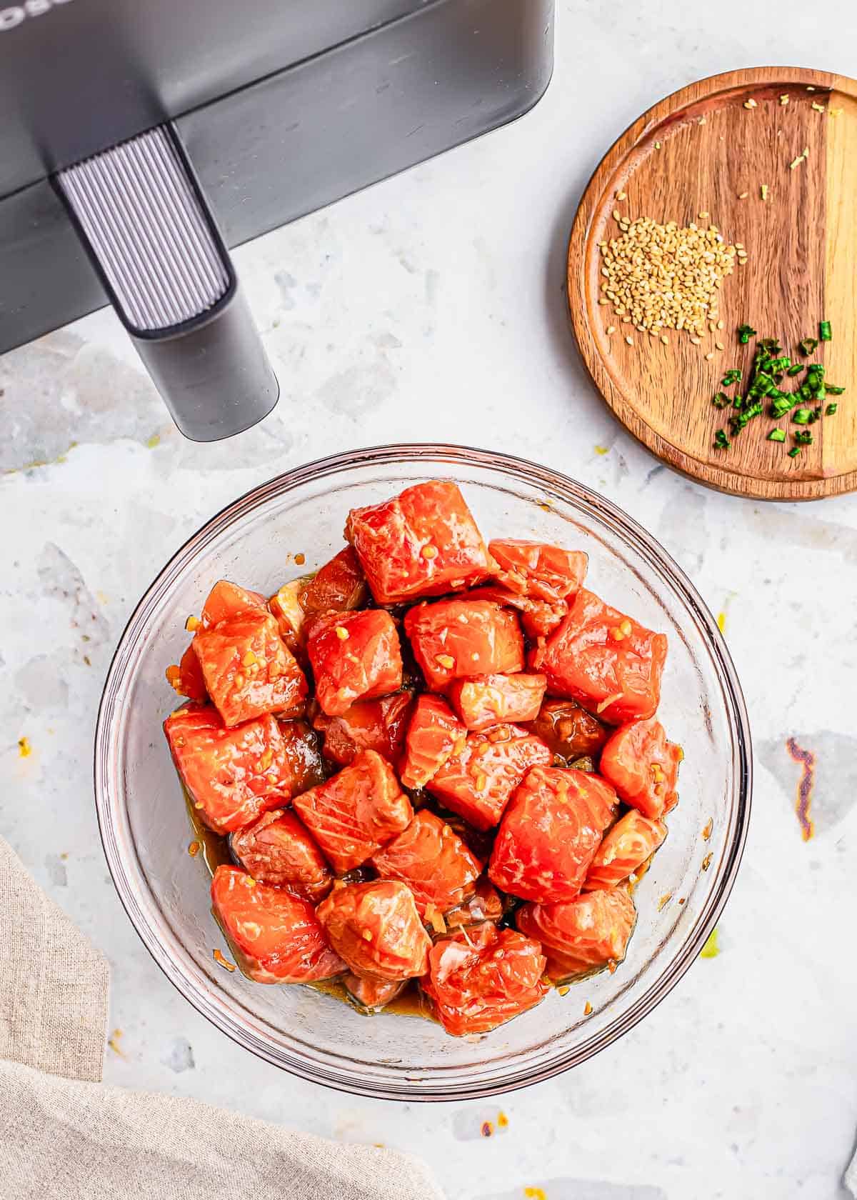 Salmon bites tossed in a marinade in a glass bowl with air fryer behind it.