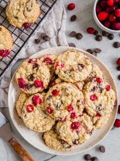 A plate of cranberry and chocolate chip cookies with chocolate chips and fresh cranberries scattered around it.