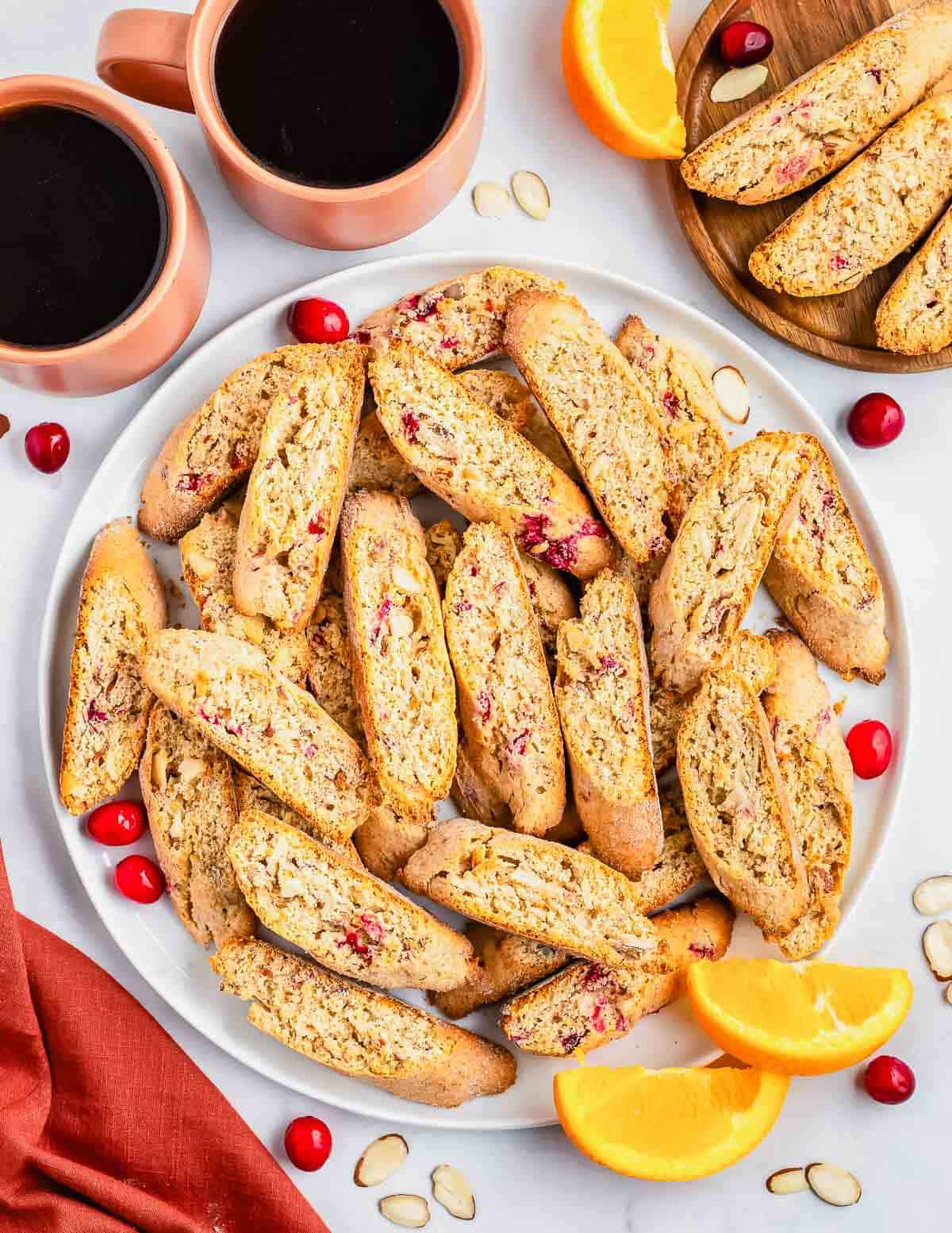 A pile of cranberry orange almond biscotti on a plate with coffee cups in the background.