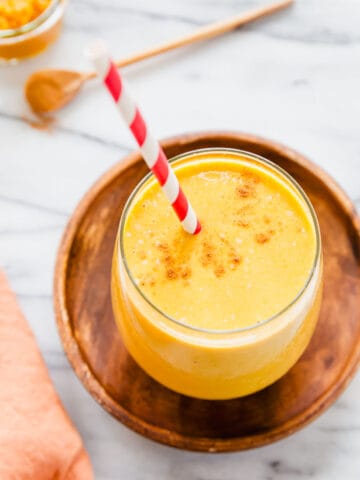 5-ingredient banana pumpkin smoothie in a glass with a red striped straw with cinnamon and pumpkin puree in the background.