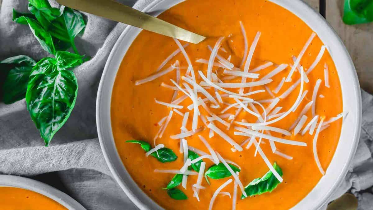 Creamy tomato basil soup garnished with grated parmesan and fresh basil in a white bowl with a gold spoon.