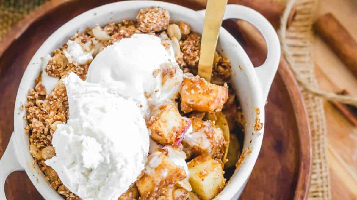 Apple crisp in a white bowl with whipped cream and cinnamon sticks.