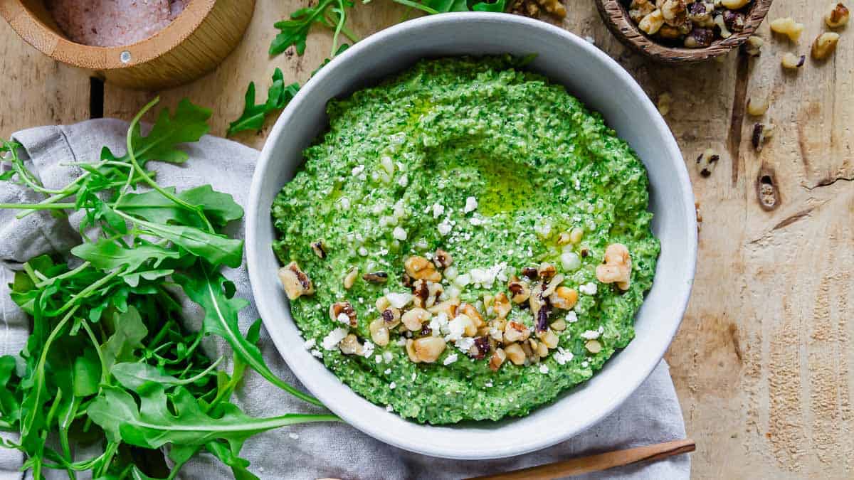 A bowl of green arugula pesto on a wooden table.