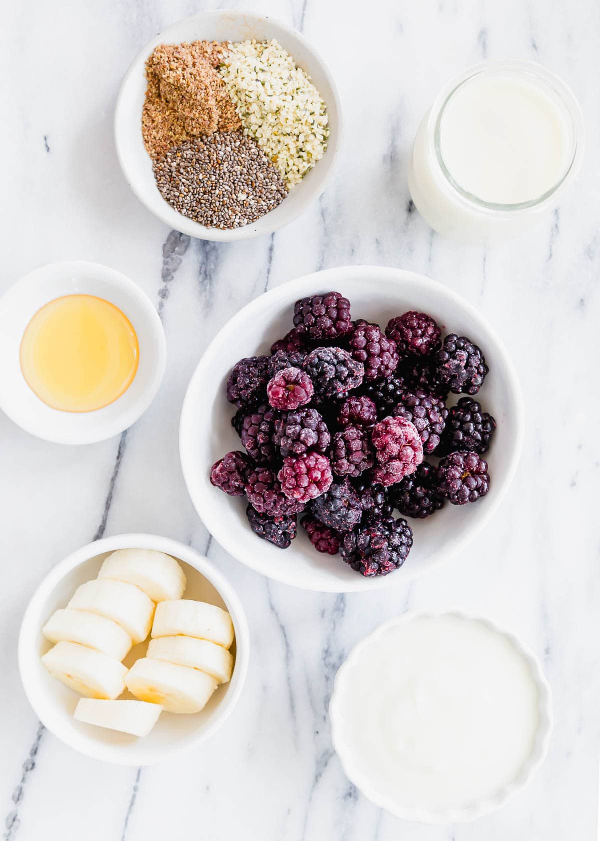 Frozen blackberries with sliced banana, honey, milk, yogurt, hemp seeds, flax and chia in bowls.