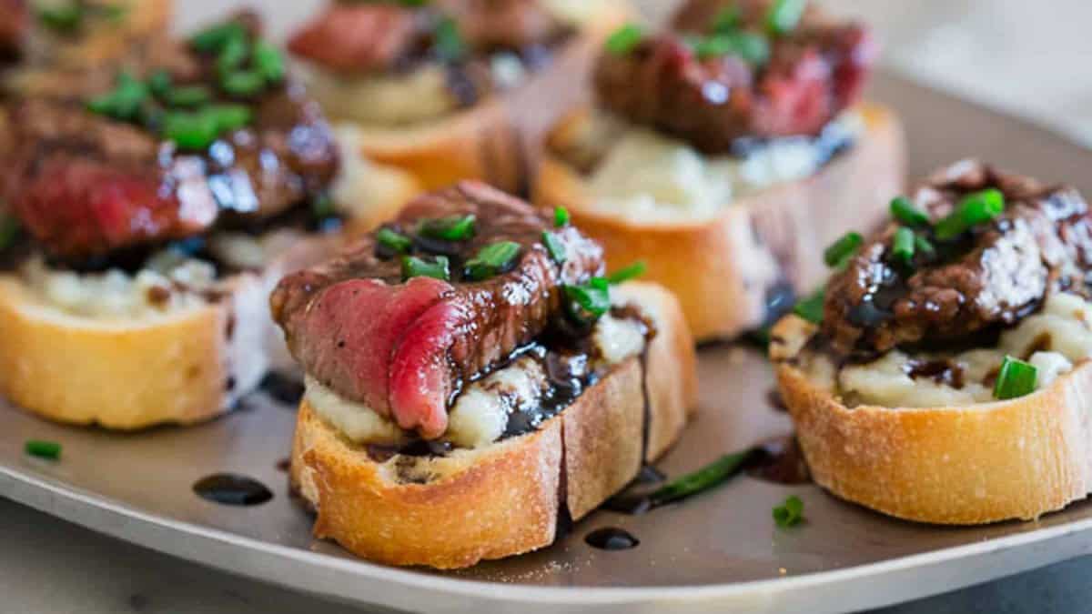 Blue cheese steak crostini on a metal platter drizzled with balsamic reduction.