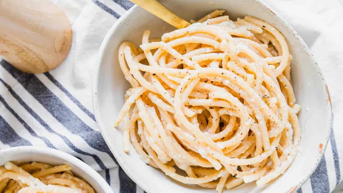 Bucatini cacio e pepe in a bowl with a gold fork and a pepper mill off to the side.