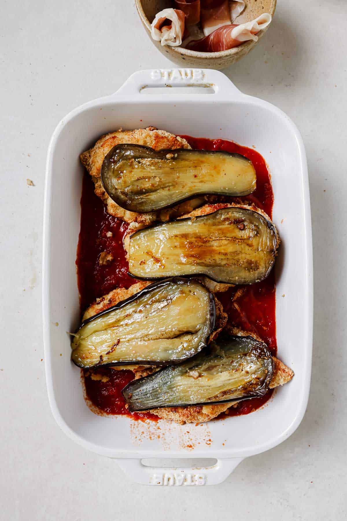 Breaded chicken topped with roasted eggplant slices in a white baking dish with sauce.