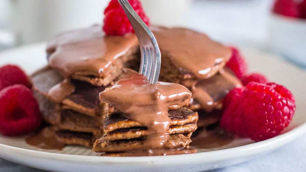 A stack of pancakes with chocolate sauce and raspberries on a plate.