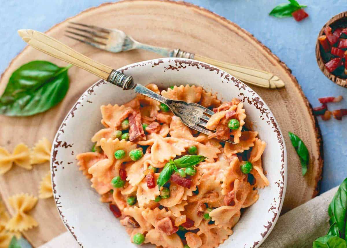 Creamy tomato farfalle with peas and prosciutto in a white bowl.