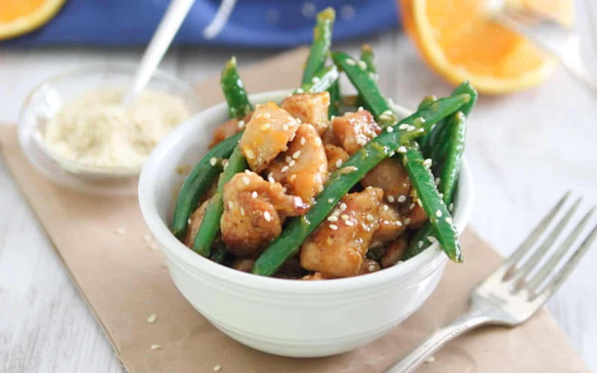 Honey orange chicken with sesame seeds and green beans in a white bowl.