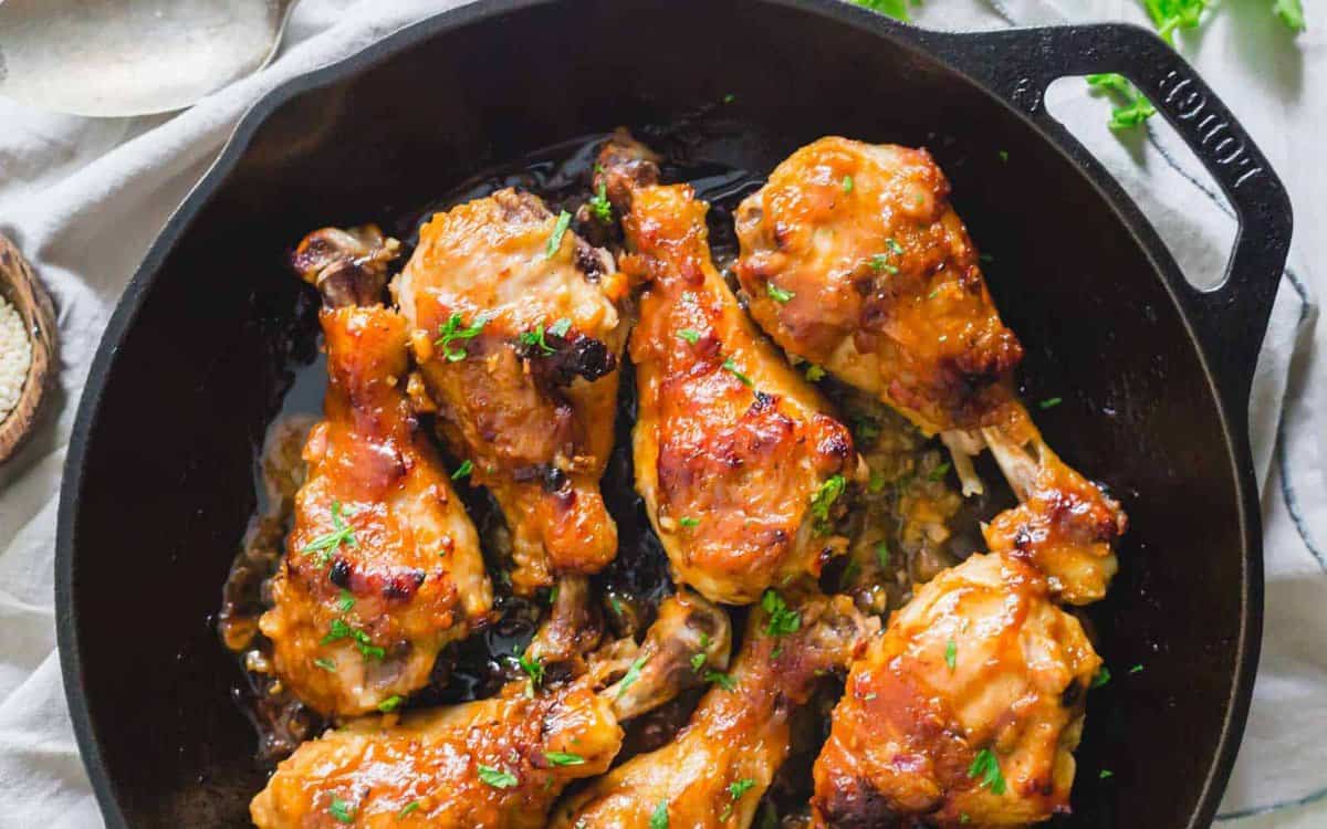 A close-up of a cast-iron skillet filled with cooked chicken drumsticks glazed with a golden, caramelized sauce, and garnished with chopped parsley. The drumsticks appear crispy and juicy, with a savory and appetizing appearance.