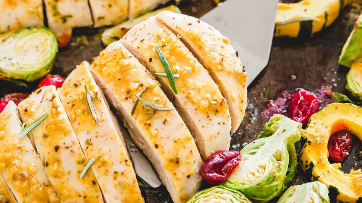 A sliced, seasoned chicken breast garnished with herbs is surrounded by roasted vegetables including brussels sprouts, cherry tomatoes, and squash on a baking sheet. A spatula is partially visible under the chicken slices.