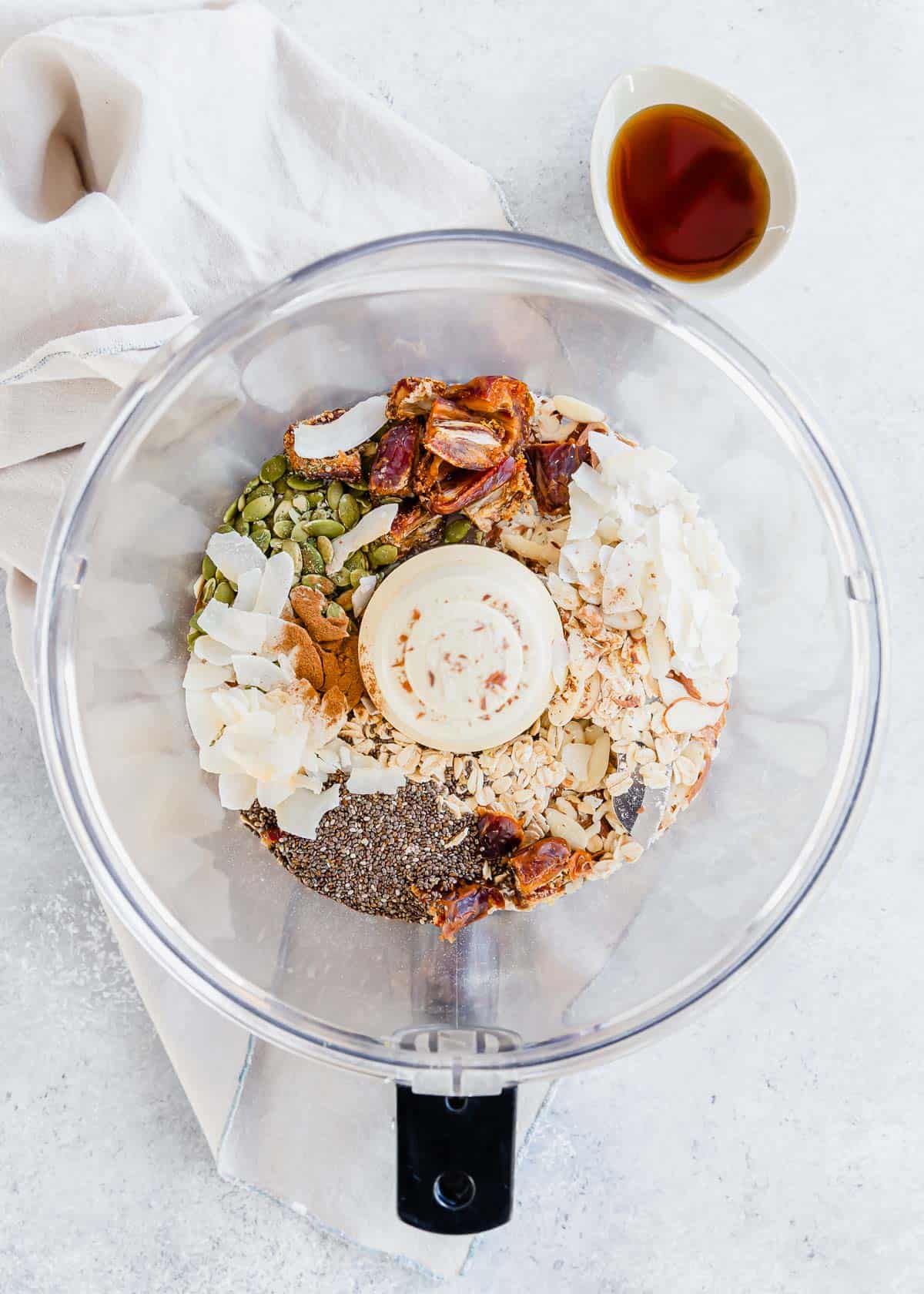 A food processor filled almonds, dates, oats, coconut, pepitas, chia seeds and a bowl of maple syrup on the side.