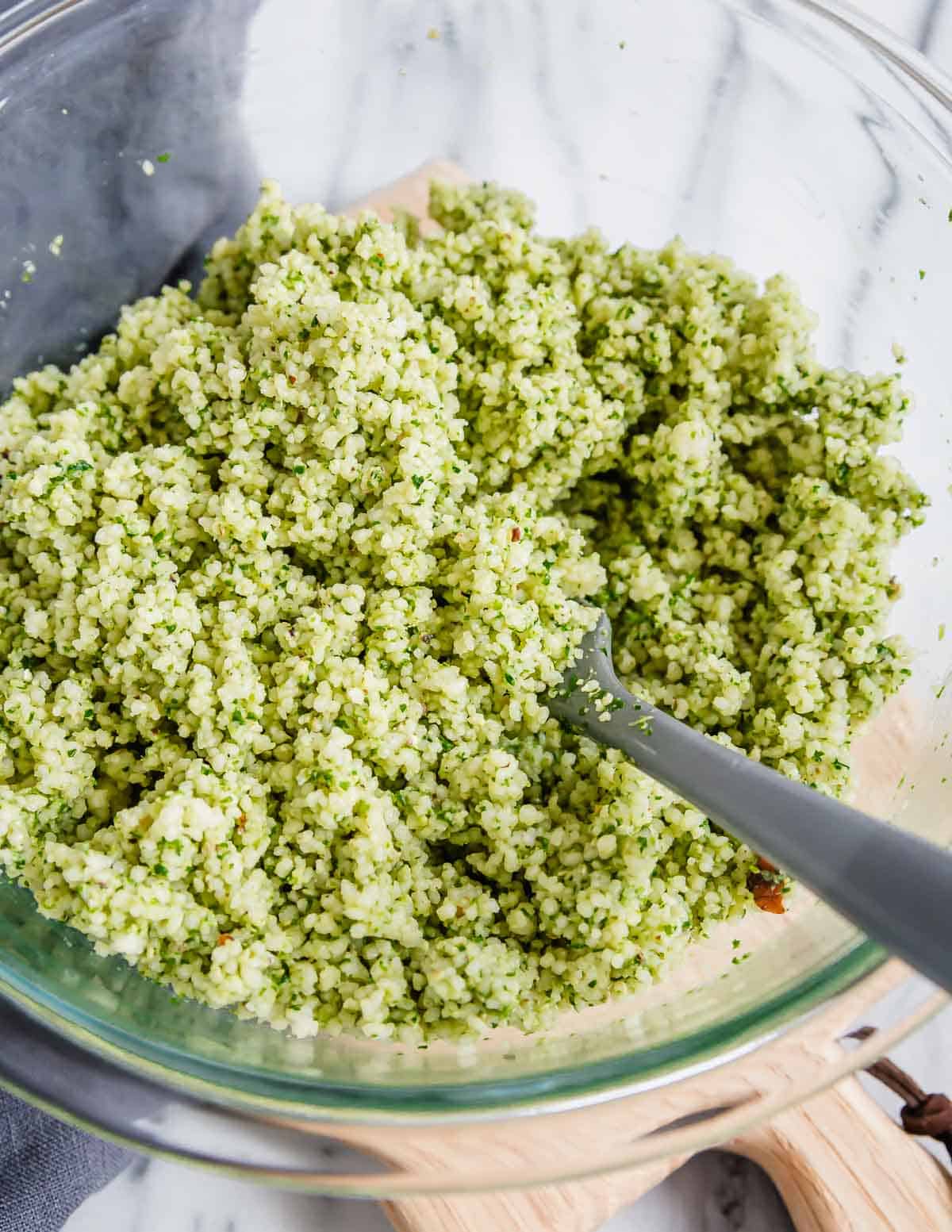 Couscous tossed with pesto in a glass bowl with a spatula.