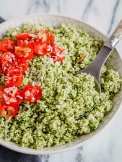 A bowl of couscous tossed with pesto with tomatoes and parmesan cheese with a serving spoon.