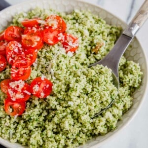 A bowl of couscous tossed with pesto with tomatoes and parmesan cheese with a serving spoon.