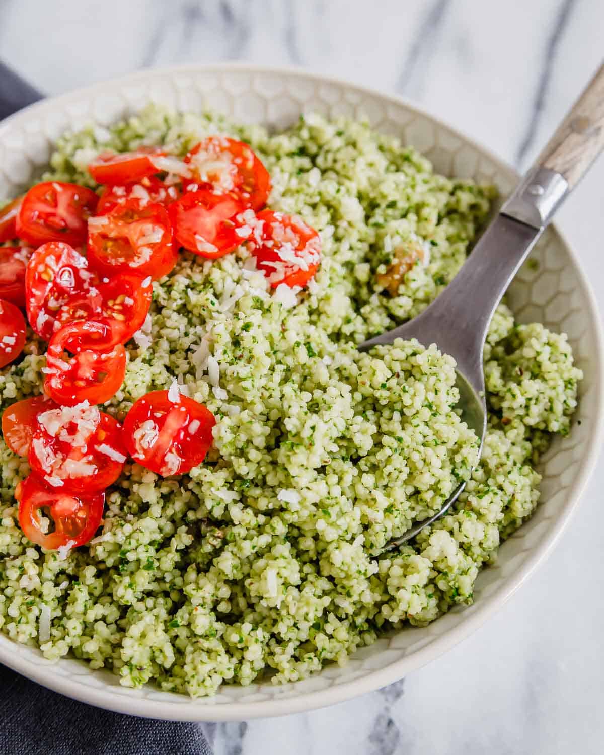 A bowl of couscous tossed with pesto with tomatoes and parmesan cheese with a serving spoon.