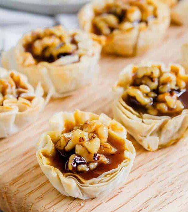 A tray of mini pumpkin pie tarts with nuts and caramel on a wooden cutting board.