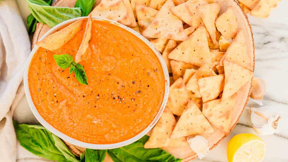 A bowl of roasted red pepper dip with crackers on a marble table.