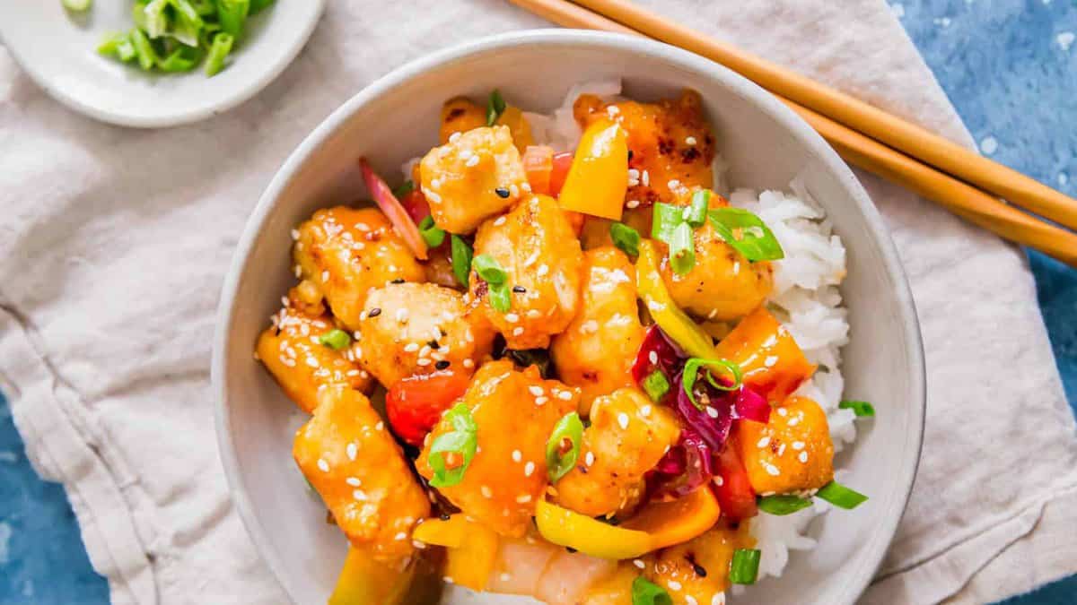 Sweet and sour tofu in a bowl with rice and chopsticks on the side.