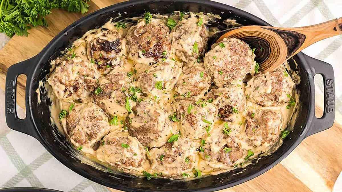 A skillet filled with meatballs and gravy on a wooden cutting board.