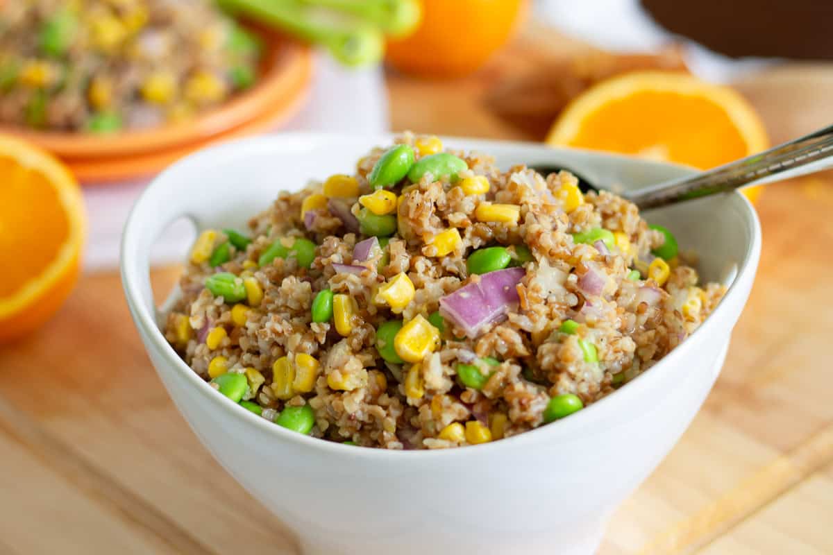 Corn, edamame and red onion bulgur wheat salad with orange dressing in a white bowl with serving spoon.