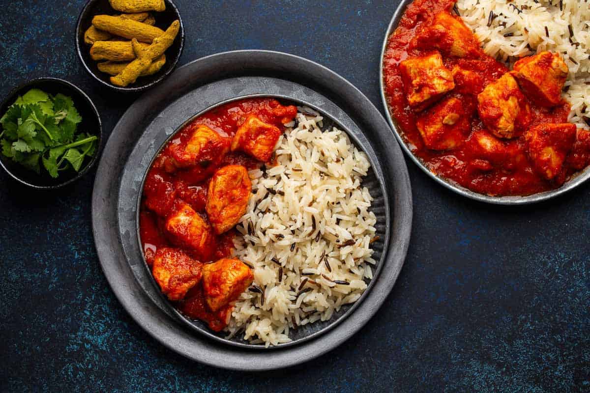 Chicken tikka masala dish with rice, flat Indian bread and spices in rustic metal plates on concrete background top view. Chicken tomato curry, turmeric root, fresh cilantro, traditional Indian meal