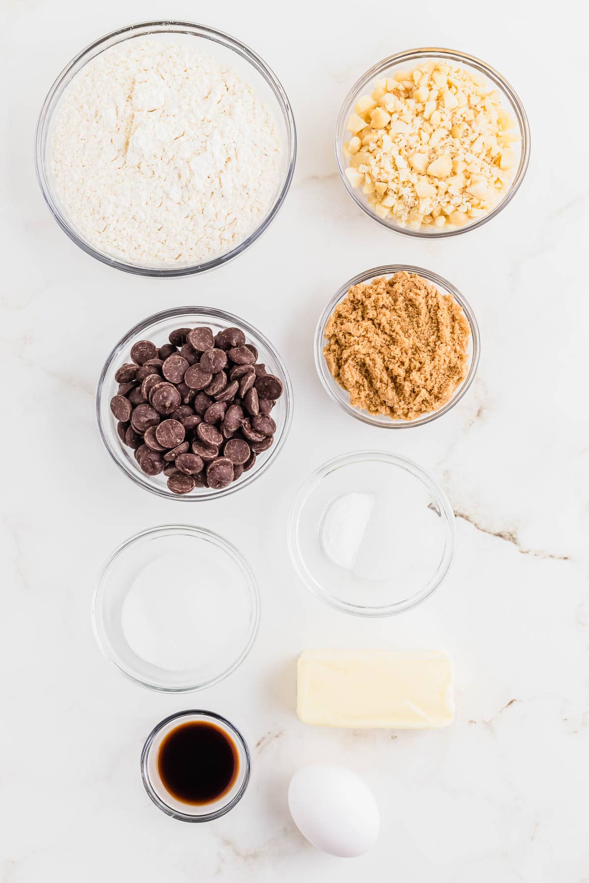 Ingredients laid out in glass bowls to make macadamia nut chocolate chip cookies on a marble surface.