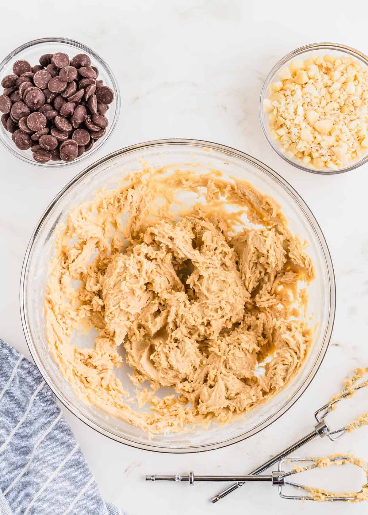 Making cookie dough in a glass bowl with chocolate chips and macadamia nuts in small bowls on the side.