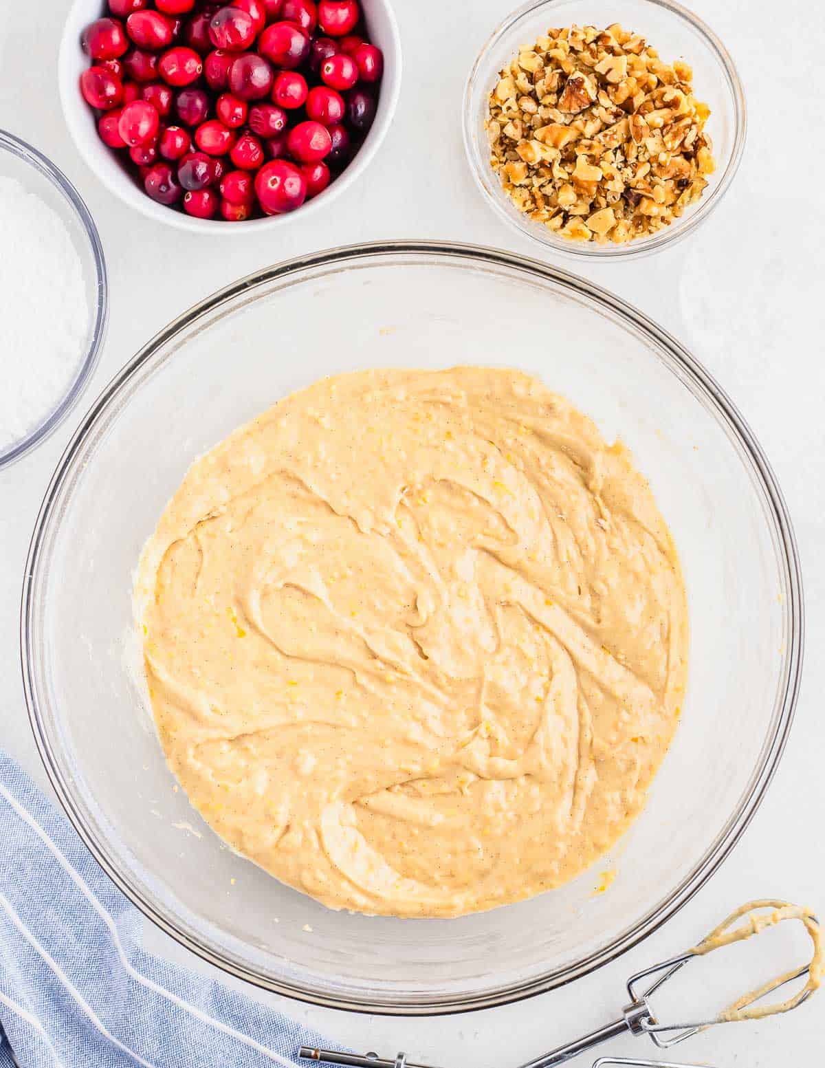 Bread batter in a large glass bowl with small bowls of fresh cranberries and chopped walnuts on the side.