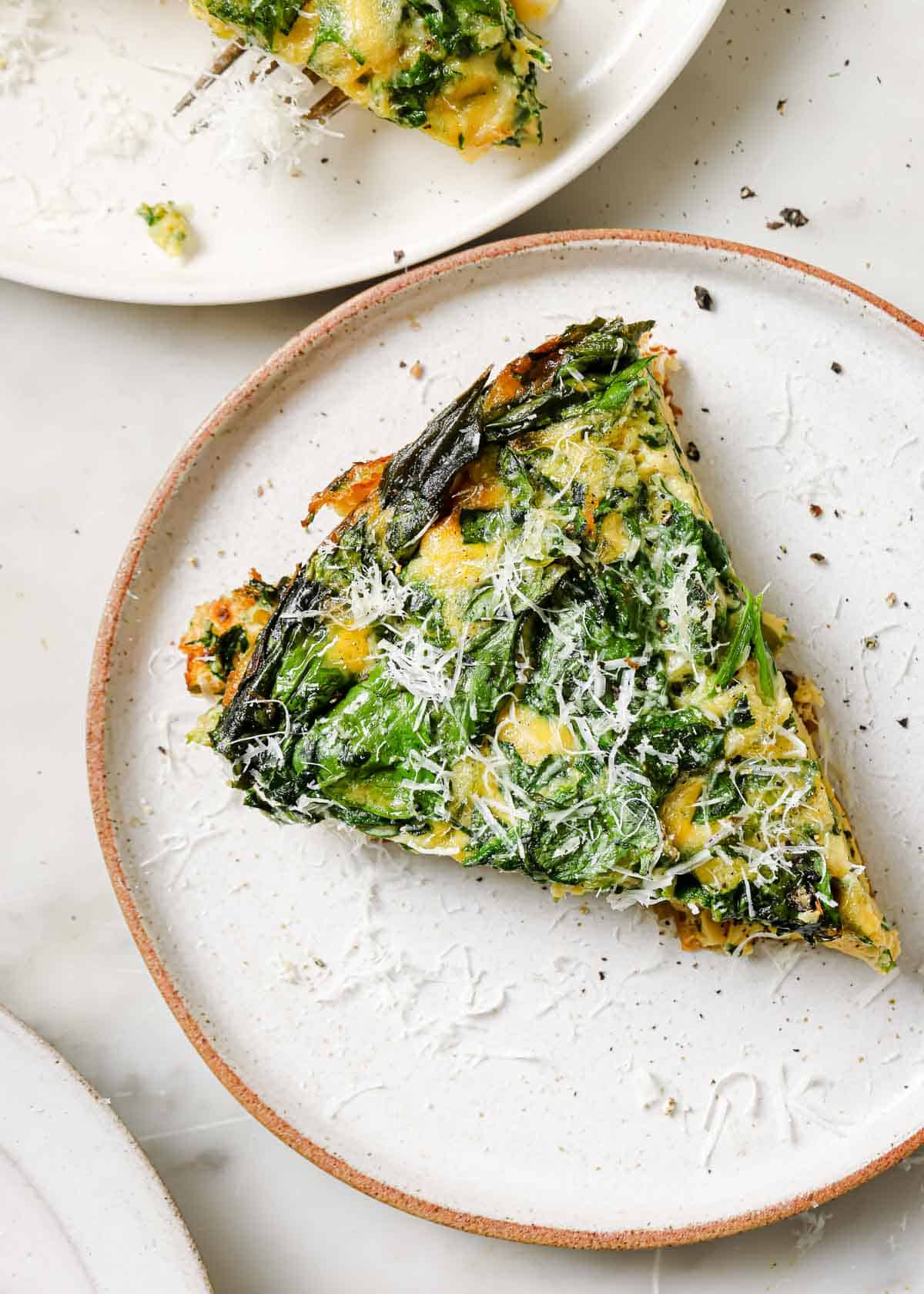 Spinach frittata slice on a white plate topped with grated parmesan cheese.
