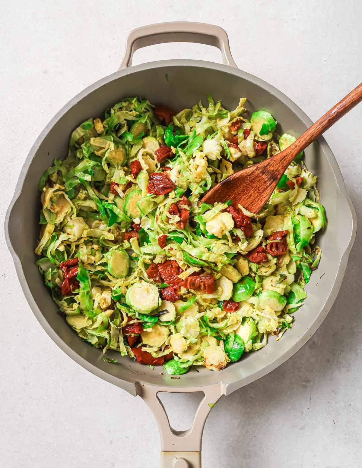 Brussels sprouts and sun-dried tomatoes in a skillet with a wooden spoon.